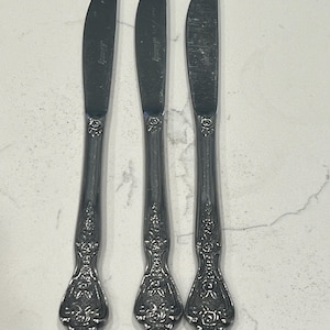 May include: Three silver butter knives with ornate floral detailing on the handles. The blades are curved and feature a subtle inscription. The knives are arranged vertically against a light-colored, textured surface.