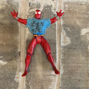 以下が含まれることがあります： 胸にクモの模様がついた赤と青のスーパーヒーローのアクションフィギュア。フィギュアは腕を伸ばし、足を広げてポーズをとっている。