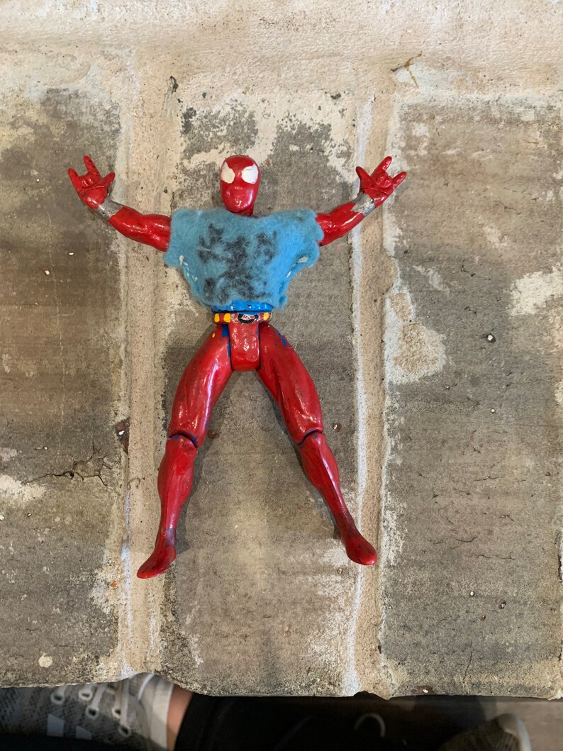 以下が含まれることがあります： 胸にクモの模様がついた赤と青のスーパーヒーローのアクションフィギュア。フィギュアは腕を伸ばし、足を広げてポーズをとっている。