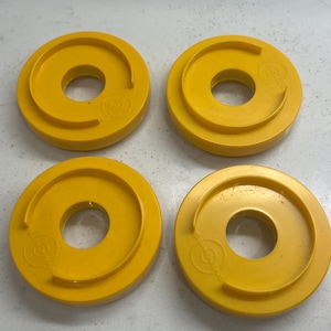 May include: Four bright yellow, circular objects with a central hole and a raised outer ring. Each object has a small, circular marking on the side. The objects are evenly spaced and arranged in a square pattern.