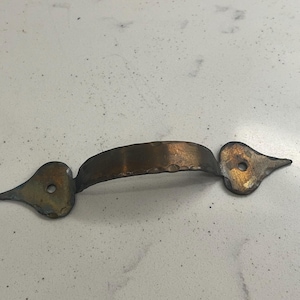 May include: A bronze-colored cabinet pull with a hammered texture on the handle. The ends are shaped like stylized bird heads, each with a drilled hole for mounting. The hardware has a rustic, aged appearance.