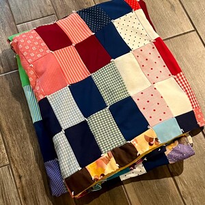 May include: Folded patchwork quilt featuring a variety of fabrics in red, blue, pink, and white. The quilt is made of squares with different patterns, including polka dots, stripes, and floral designs. The quilt is folded and appears to be handmade.