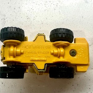 Puede incluir: Un coche de juguete amarillo Hot Wheels 3 Wheel Loader con neum&aacute;ticos negros. La parte inferior del juguete es visible, mostrando el logotipo de Hot Wheels y otro texto. El juguete est&aacute; hecho de metal y pl&aacute;stico.