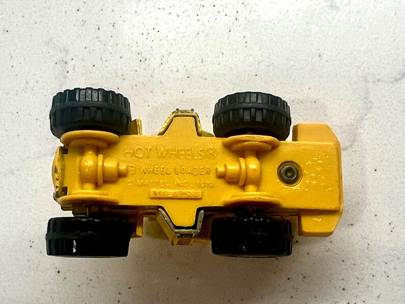 Puede incluir: Un coche de juguete amarillo Hot Wheels 3 Wheel Loader con neum&aacute;ticos negros. La parte inferior del juguete es visible, mostrando el logotipo de Hot Wheels y otro texto. El juguete est&aacute; hecho de metal y pl&aacute;stico.