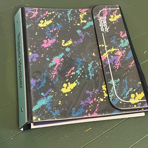 May include: A black Trapper Keeper binder featuring a colorful paint splatter pattern in shades of pink, yellow, teal, and purple. The binder includes a vertical flap closure with the Trapper Keeper logo and a light blue spine with the brand name.