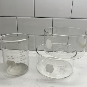 May include: Two clear glass beakers. The beaker on the left has a pouring spout and measurement markings up to 300 ml. The beaker on the right has a cylindrical shape with the text "125 X 65 KIMAX" printed on it. Both beakers are empty.
