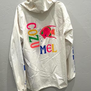 May include: An off-white long-sleeve hooded pullover featuring a graphic of a pink and green tropical fish. The text COZUMEL is printed in large, colorful letters across the back, with blue and yellow accents on the sleeves and a drawstring hood.