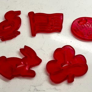 May include: Five red plastic cookie cutters in various shapes: a duck, a flag, an egg with "Happy Easter" text, a rabbit, and a shamrock. These cutters are likely used for baking themed cookies.