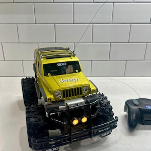 May include: Yellow remote-controlled Jeep toy with black accents. The toy has large, rugged tires and the word "Jeep" is visible on the front. The brand name "Nikko" is printed on the front bumper and the remote control.