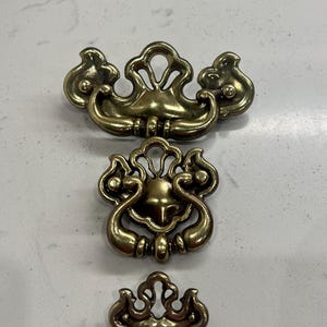 May include: Three antique brass cabinet pulls with ornate designs. The pulls are different sizes and have a vintage look.