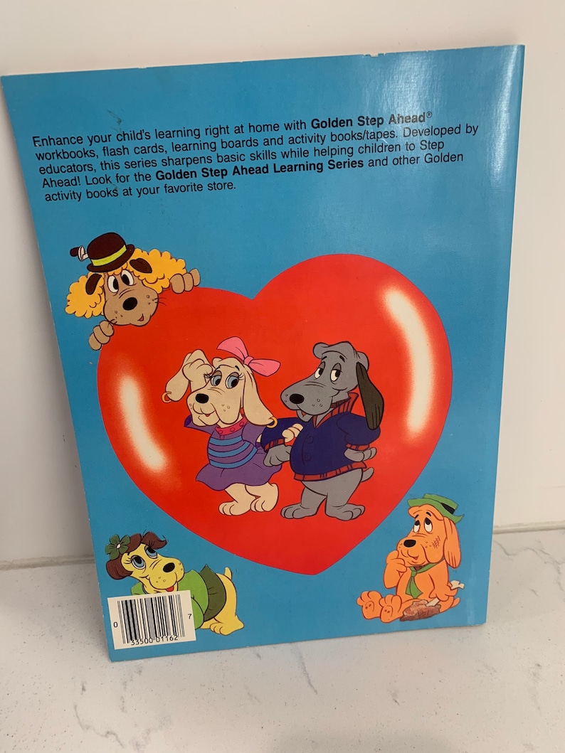 May include: A book cover with a blue background and a large red heart. The heart has four cartoon dogs on it. The text on the cover reads "Golden Step Ahead" and "Golden Step Ahead Learning Series".