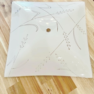 Può includere: Plafoniera quadrata in vetro bianco con un design a forma di grano in oro e grigio. La plafoniera ha un elemento centrale circolare ed è posta su una superficie di legno.