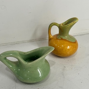 May include: Two small ceramic pitchers. One is light green, the other is orange with a green top. Both have a handle and a spout. The pitchers are likely for decorative purposes or for serving small amounts of liquid.