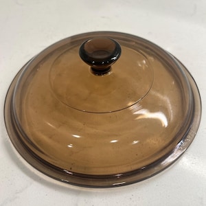May include: A round, transparent amber-colored glass lid featuring a central knob handle. The lid has a smooth, curved surface and a slightly raised rim. Embossed text is visible along the outer edge of the glass, indicating the product details.