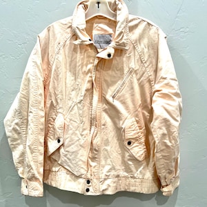 May include: A light peach-colored jacket with a zip-up front, two front pockets, and a stand-up collar. The jacket has a label on the collar and is hanging on a white hanger. The jacket is made of a lightweight material.
