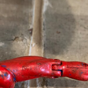 以下が含まれることがあります： 赤いプラスチック製のフィギュアの腕のクローズアップ。青いストライプが入っています。腕は肘で曲がっていて、フィギュアの著作権情報が確認できます: &copy; 1991 Marvel.