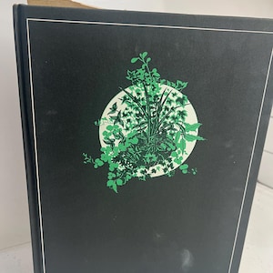 May include: A hardcover book with a black cover featuring a white border and a circular design of green foliage. The design is centered on the cover, with a variety of leaves and plants.