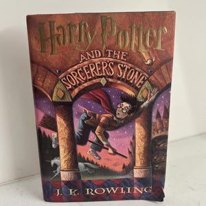 May include: A hardcover book with a red, brown, and gold cover. The cover features an illustration of Harry Potter flying on a broomstick with a unicorn in the foreground. The title of the book is "Harry Potter and the Sorcerer's Stone" with the author's name "J.K. Rowling" at the bottom.