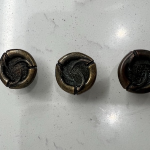 Peut inclure: Trois boutons de meuble anciens en bronze avec un motif en spirale. Chaque bouton est rond avec un centre texturé en relief et une finition bronze foncé, pour la restauration de meubles.