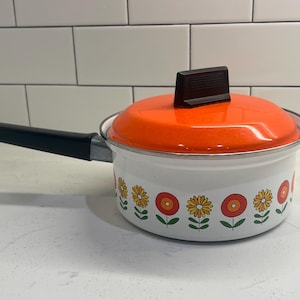 Puede incluir: Un cazo de metal esmaltado blanco con una tapa de color naranja brillante y un mango negro. El exterior de la olla presenta un patrón repetitivo de flores amarillas y naranjas con tallos verdes. Un pomo negro está centrado en la tapa.