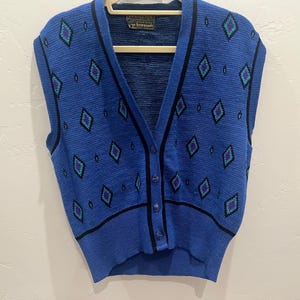 May include: A blue knit sweater vest featuring a V-neckline, a button-front closure, and a repeating diamond pattern in black, teal, and purple. The garment has a ribbed hem and armholes, with a Pendleton brand label visible at the inner neckline.