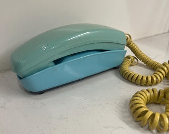 Könnte beinhalten: Ein zweifarbiges Retro-Telefon in Hellblau und Türkis. Der Hörer liegt auf einer Basis, mit einem gewendelten gelben Kabel, das sich vom Telefon erstreckt. Das Telefon hat ein Retro-Design, wahrscheinlich aus der Mitte des 20. Jahrhunderts.
