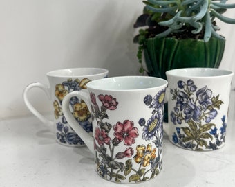 May include: Three white ceramic mugs with floral patterns. The mugs have a white background with colorful flowers and leaves. The mugs are arranged in a row on a white surface.