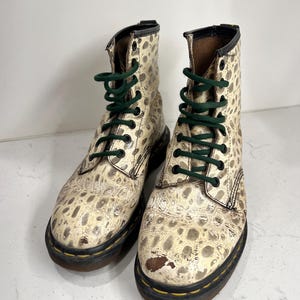 Puede incluir: Un par de botas de cuero blanco con un patrón de manchas blancas y negras. Las botas tienen cordones verdes y una suela negra.