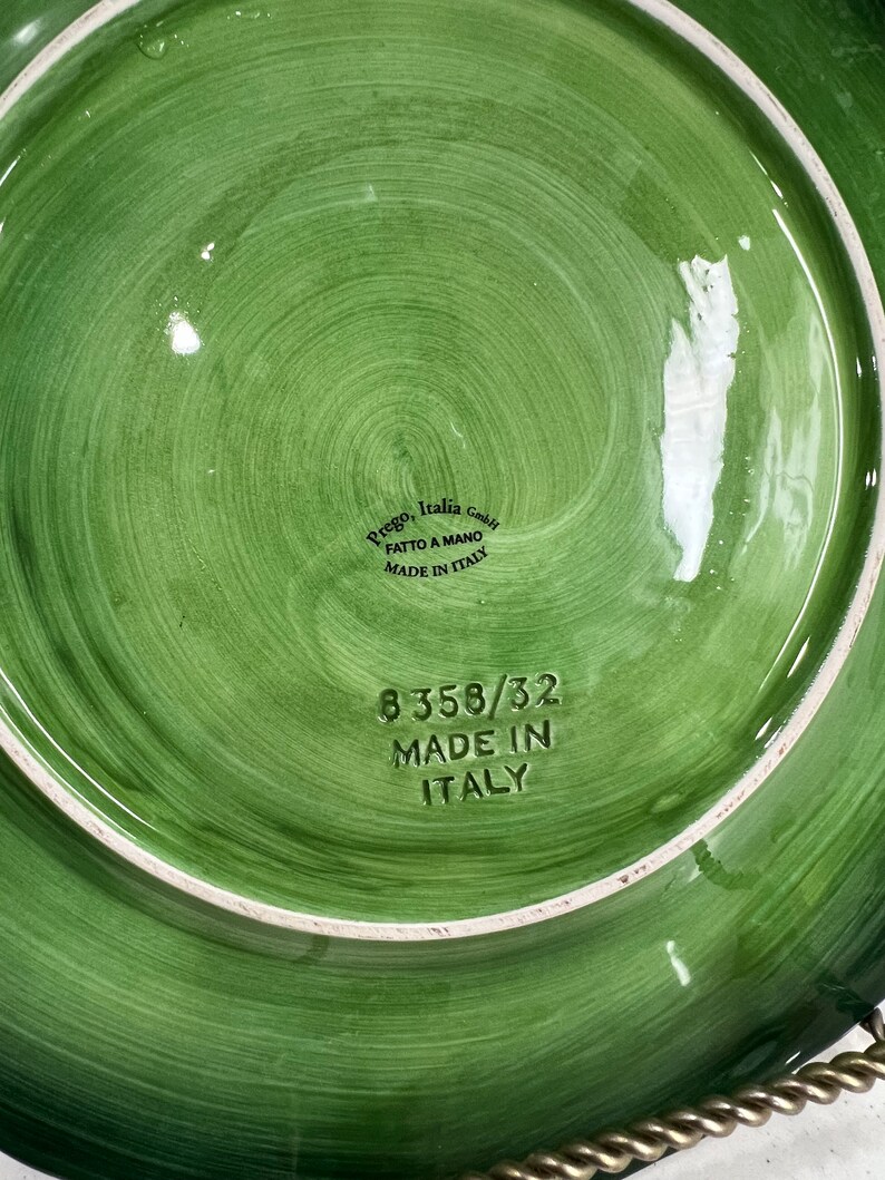 Pu&ograve; includere: Piatto in ceramica verde con bordo bianco e design circolare. Il fondo del piatto ha il testo "Prego Italia GmbH" e "Fatto a Mano Made in Italy" in nero. Il piatto ha anche il numero "8358/32 Made in Italy" in nero.