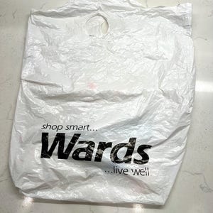 Op de afbeelding: Een gekreukelde witte plastic boodschappentas met een rond handvatgat. De tas heeft de tekst "shop smart... Wards ...live well" in zwarte letters.