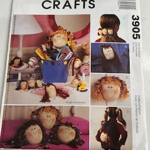 May include: A McCall's Crafts pattern envelope featuring various doll designs. The cover showcases doll faces with yarn hair, displayed in different ways, including hair accessories and a denim jacket. The text "McCall's Crafts" is at the top.