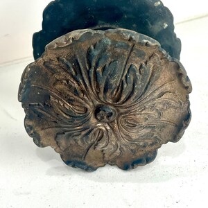May include: A round, metallic door knob featuring a dark, weathered bronze finish and an intricate, raised floral or leaf-like pattern radiating from the center. The knob has a scalloped edge and a deep, dark patina within the recessed areas.