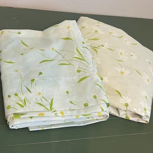 May include: Two folded pillowcases featuring a light blue and pale green background with a repeating pattern of white daisy flowers, thin green stems, and small yellow centers. The fabric displays a soft, floral botanical print throughout.
