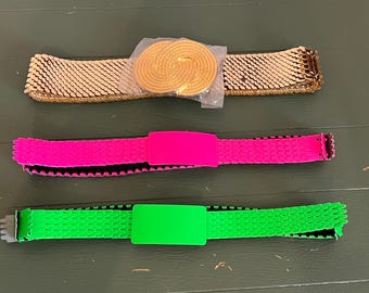 Op de afbeelding: Drie riemen zijn horizontaal weergegeven. De bovenste riem is goudkleurig met een metalen, getextureerd schakelontwerp en een ronde gouden gesp die gedeeltelijk is bedekt met plastic. De middelste riem is neonroze met een rechthoekige gesp. De onderste riem is neongroen.
