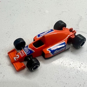 Könnte beinhalten: Oranges Spielzeug-Rennwagen mit schwarzen Rädern. Das Auto hat blaue und weiße Akzente mit der Nummer 2 auf der Vorderseite. Die Wörter "Racing" und "MIC" sind auf dem Auto sichtbar.