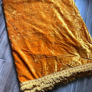 May include: A rectangular mustard yellow velvet throw blanket featuring a crushed texture and a decorative trim along the bottom edge. The trim consists of a woven band with a repeating geometric pattern and a dense fringe of matching tassels.
