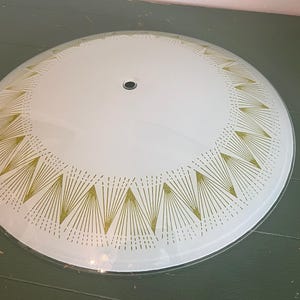 以下が含まれることがあります： オリーブグリーンの放射状の幾何学模様が縁に施された、円形の白いガラス製シーリングライトシェード。シェードの中央は無地の白で、取り付け用の円形の穴が一つあり、縁は透明なガラスになっています。