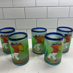 Puede incluir: Juego de cinco vasos de chupito de vidrio esmerilado con borde azul. Cada vaso presenta una colorida ilustración de una persona que lleva una cesta. La persona lleva un sombrero, una camisa roja y pantalones blancos. El fondo es verde.