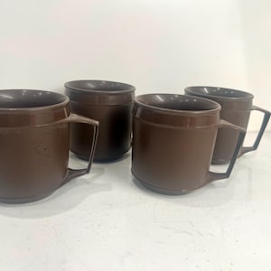 May include: A set of four dark brown plastic mugs, each featuring a cylindrical body, a wide rim, and a geometric angular handle. The mugs are arranged in a group, displaying a uniform solid color and a smooth, matte finish throughout their design.