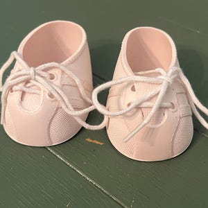 May include: A pair of light pink doll trainers featuring a textured fabric-like finish, a rounded toe cap, and white laces tied in a bow. The shoes have a low-top design with visible eyelets and side stripes, constructed from a moulded material.