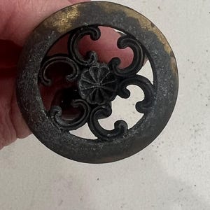 Könnte beinhalten: Ein runder, messingfarbener Schrank-Knopf mit einem dekorativen, durchbrochenen Design. Der Knopf hat eine dunkle Patina und eine leicht abgenutzte Oberfläche.