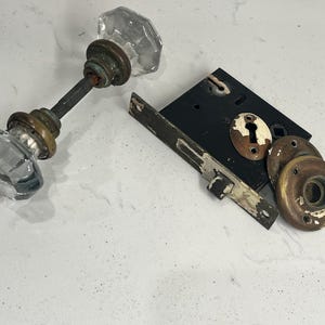 May include: A set of two clear, faceted glass door knobs connected by a metal spindle, alongside a black rectangular mortise lock box with a worn brass keyhole plate and two circular brass rosettes showing signs of age and paint wear.