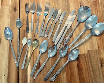 May include: An assortment of vintage silverware, including forks, knives, and spoons, arranged on a wooden surface. The silverware features various designs and sizes, with some pieces showing ornate detailing. The collection includes serving spoons and butter knives.