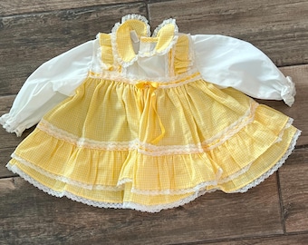 May include: A vintage baby dress featuring a yellow and white gingham pattern skirt with white lace trim. The dress has a white top with long sleeves and a ruffled collar. A yellow ribbon bow accents the waist.