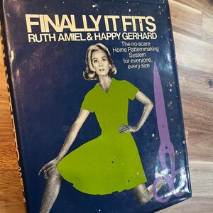 Op de afbeelding: Een blauwe boekomslag met een vrouw in een groene jurk en een schaar. De titel van het boek is "Finally It Fits" door Ruth Amiel & Happy Gerhard. De tekst onder de titel luidt "The no-scare Home Patternmaking System for everyone, every size."