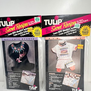 May include: Two packages of Tulip Secret Recipes fabric painting kits. Each package includes a stencil, transfer paper, and fabric paint. The first package features a black dress with a delicate heart design. The second package features a white t-shirt with the word "SURF" printed on it.