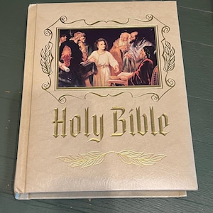 May include: A beige hardcover Holy Bible with gold lettering and a gold-outlined rectangular illustration on the front cover. The illustration depicts a group of figures in historical clothing. A gold leaf design is printed below the title.