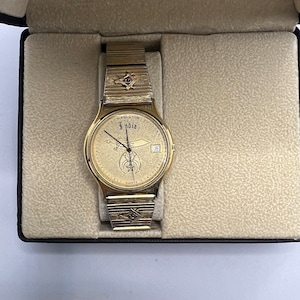 May include: A gold-toned wristwatch with a white face and black Roman numeral hour markers. The watch has a gold-toned band with a Masonic symbol on the clasp. The watch is in a black box with a beige interior.