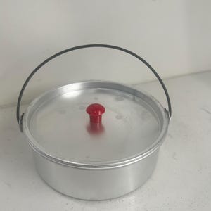 Pode incluir: Uma panela de cozinha redonda de alumínio com tampa e alça preta. A tampa tem um botão vermelho no centro. A panela está sobre uma superfície branca.
