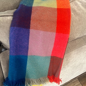May include: A colorful, rectangular blanket with a patchwork design. The blanket features squares of blue, purple, pink, green, orange, and red. The bottom edge has a fringe of various colors. The blanket is draped over a light gray couch.